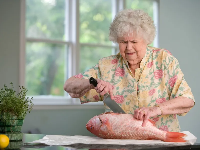 Mujer mayor preparando un pescado sobre la encimera de su cocina junto a una ventana